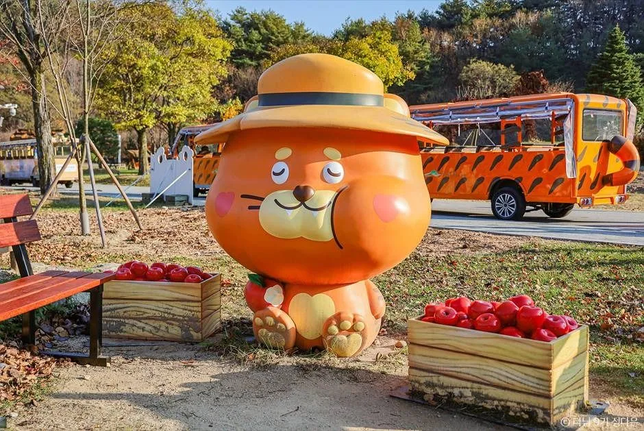 모자를 쓴 주황색 곰 모양 조형물과 사과 상자 뒤로 호랑이 무늬를 칠한 트램이 지나가는 백두대간수목원