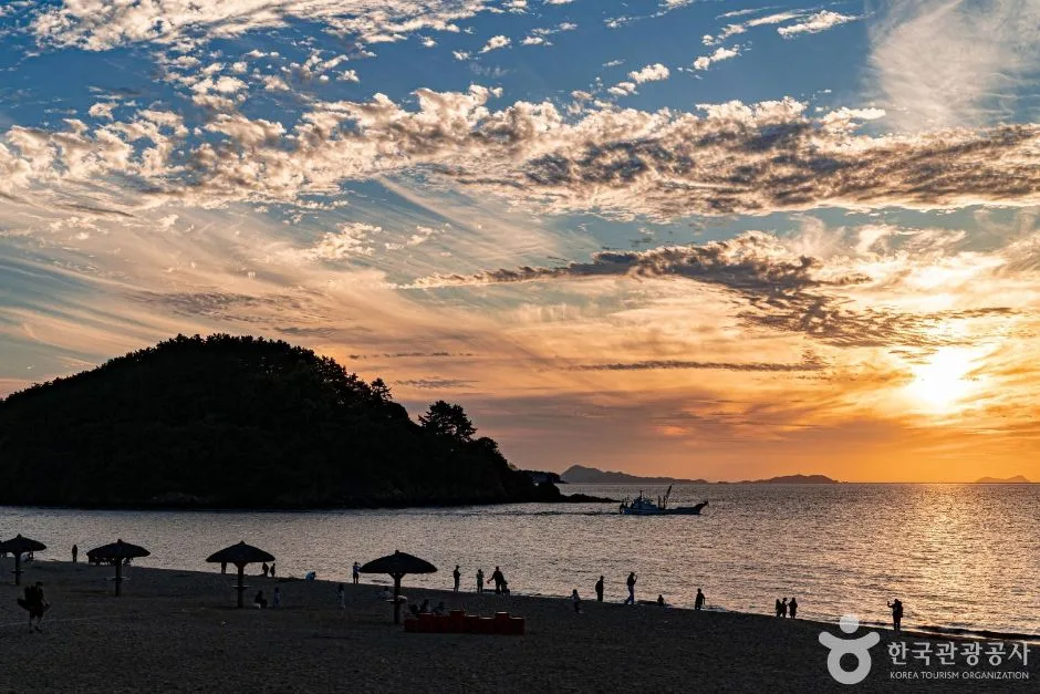 해가 지는 변산해수욕장, 금빛 하늘 아래 바다로 솟은 작은 섬과 모래사장에 늘어선 파라솔, 물가를 걷는 사람들