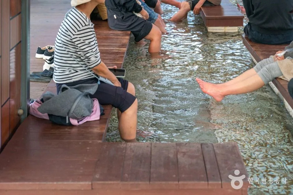 나무 의자에 나란히 앉은 사람들이 맑은 온천수가 흐르는 족욕탕에 발을 담그고 있는 수안보 족욕 체험장