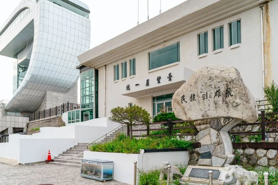 통일전망대 글씨가 걸린 하얀 건물 앞 화단에 큰 표지석이 놓이고 옆으로 곡선 유리 타워가 솟은 모습